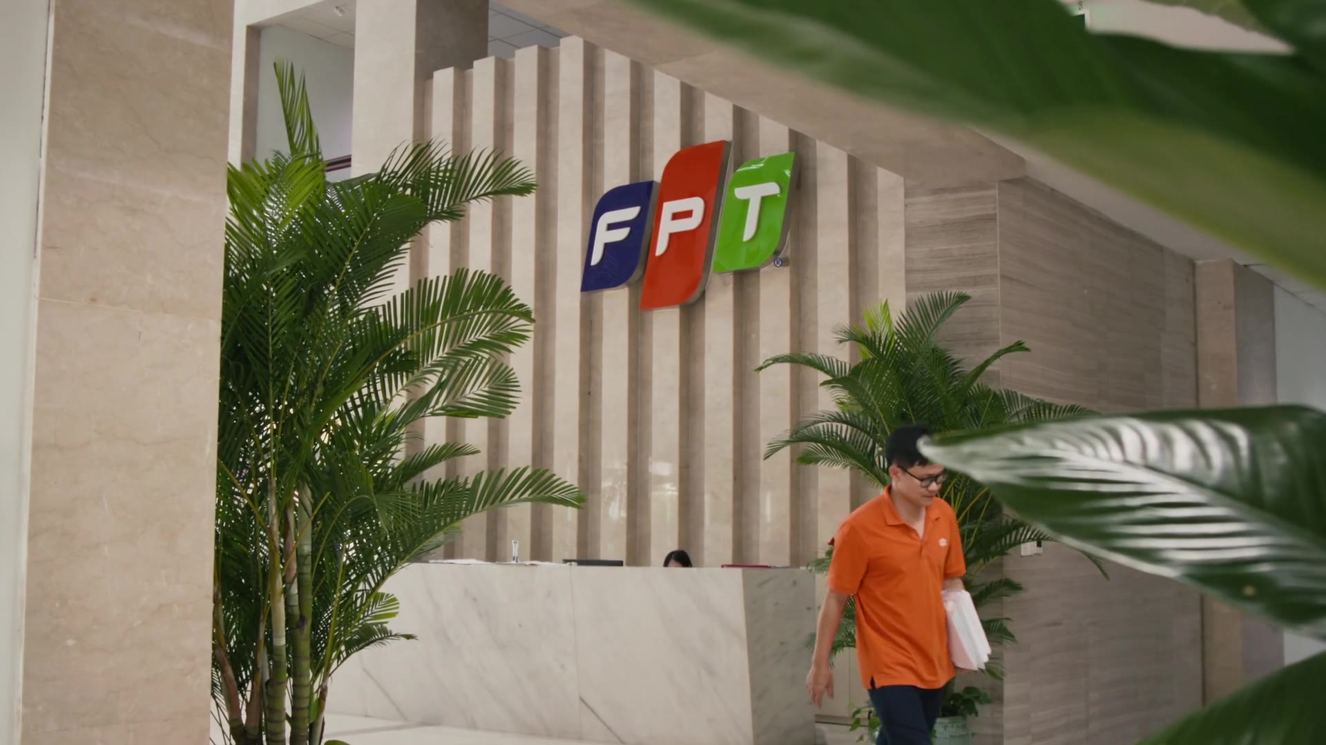 An FPT Telecom employee walking past the FPT logo in the office reception, framed through leaves